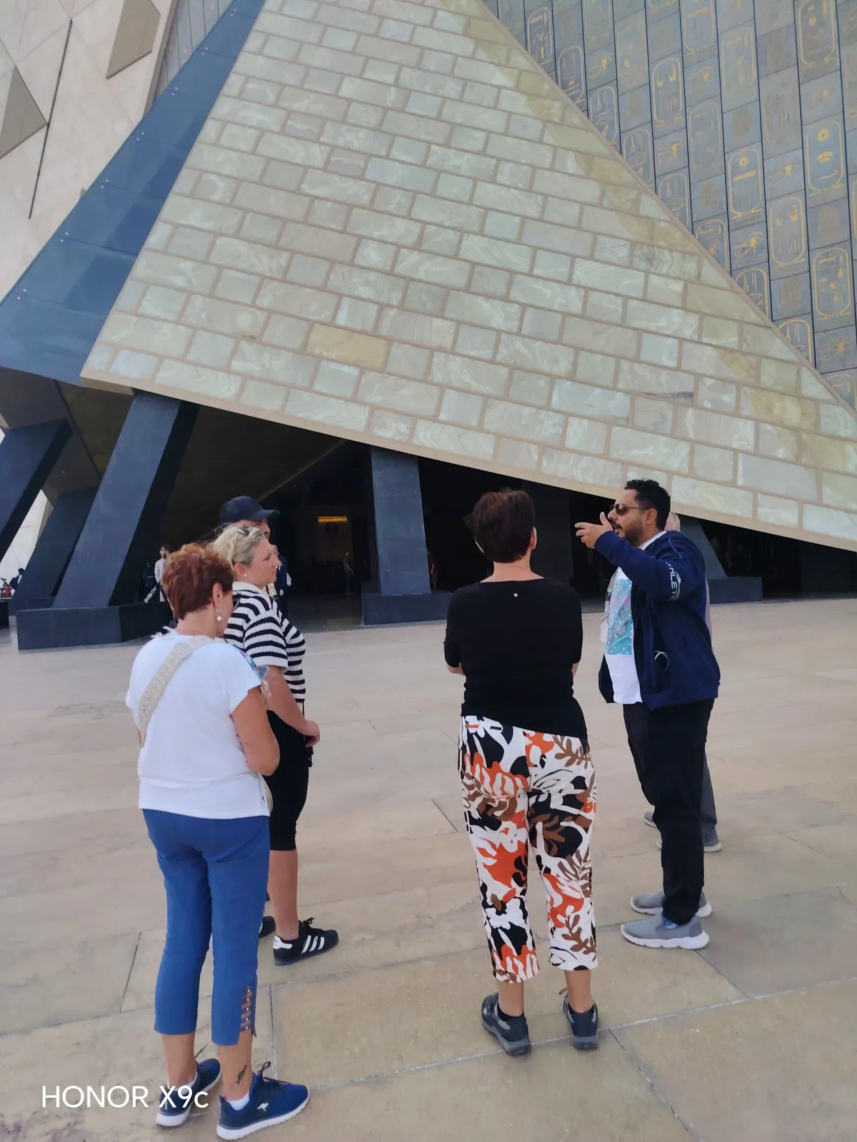 Au&szlig;enansicht des Grand Egyptian Museum mit der pyramidenf&ouml;rmigen Fassade und Besuchergruppe