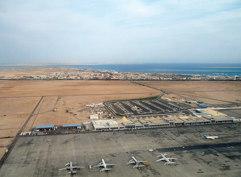 Luftaufnahme des Flughafens Hurghada mit Terminal, Flugzeugen und K&uuml;stenstadt am Roten Meer im Hintergrund.