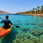 Kajakfahrer paddelt im t&uuml;rkisfarbenen Wasser vor der K&uuml;ste von Hurghada mit Palmen und Bergen im Hintergrund.