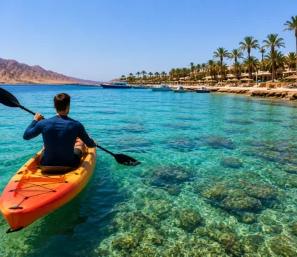Kajakfahrer paddelt im t&uuml;rkisfarbenen Wasser vor der K&uuml;ste von Hurghada mit Palmen und Bergen im Hintergrund.